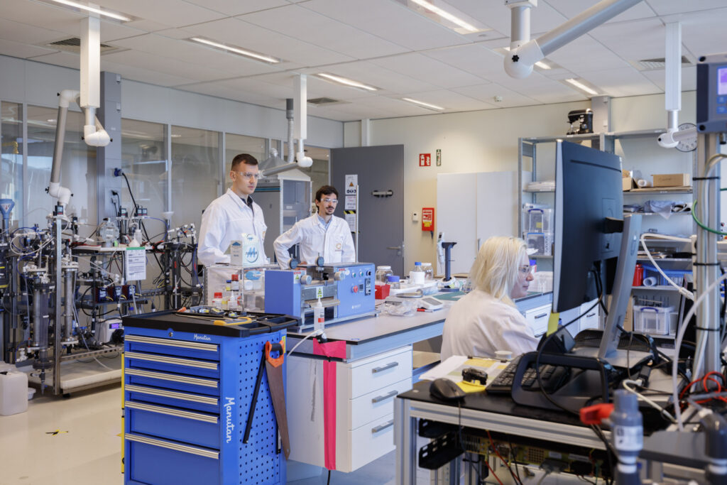 View of a laboratory with two standing men and a sitting woman. The two men are behind a blue lab bench with a lot of machines and lab equipment. In front of the bench is the woman looking at a computer screen.
