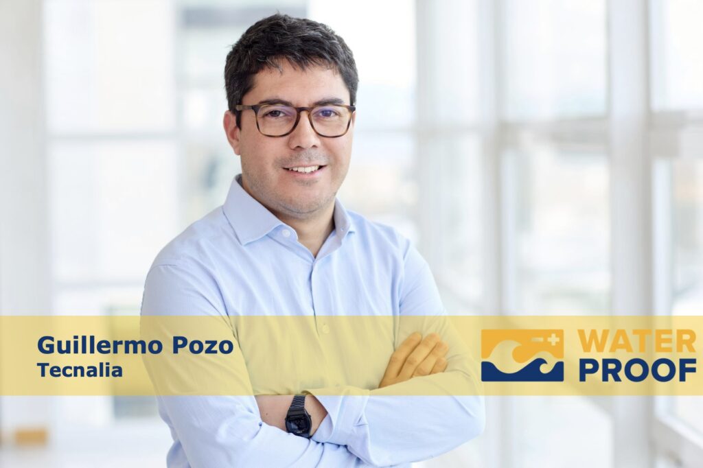 Portrait photo of Guillermo Pozo from Tecnalia. He is a dark-haired man around 40, with glasses short dark hair and a light blue shirt. The background is very bright with blurry windows.
