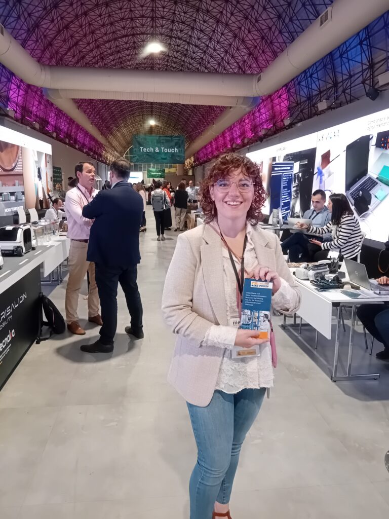 Maddalena Bettoni at meetechSpain 2024. She is a young woman with curly medium-long, brown hear wearing a white blazer and jeans. She is standing in an exhibition hall with a curved ceiling and holds a brochure of the WaterProof project.